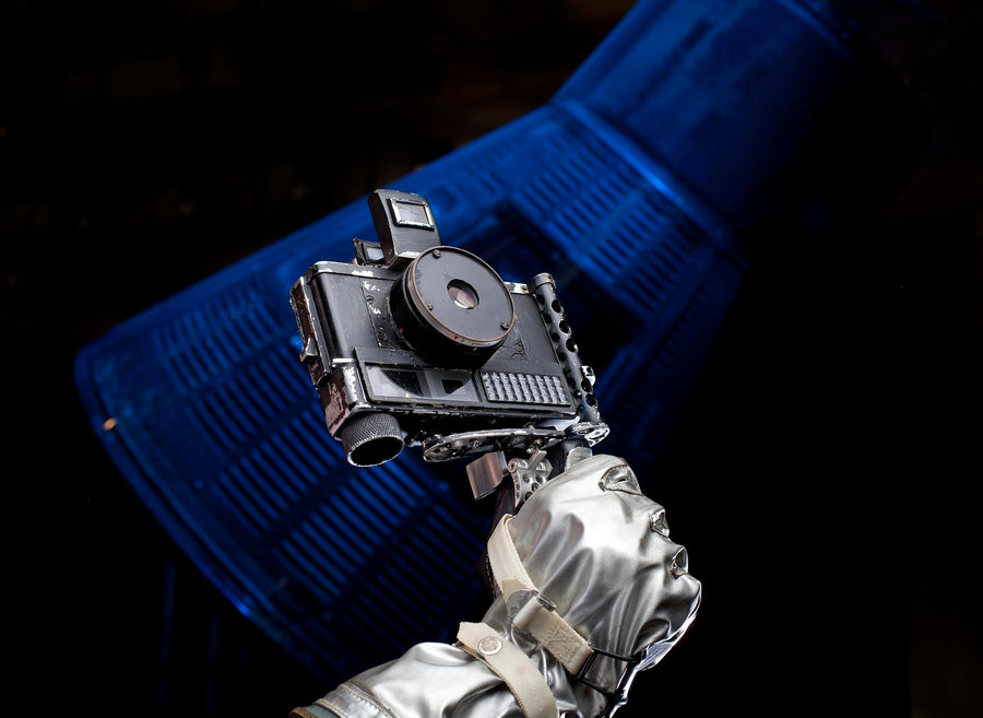 John Glenn's Ansco Autoset camera from Friendship 7, 1962, on its pistol grip and held in a silver spacesuit glove, with a spacecraft lit in blue behind it