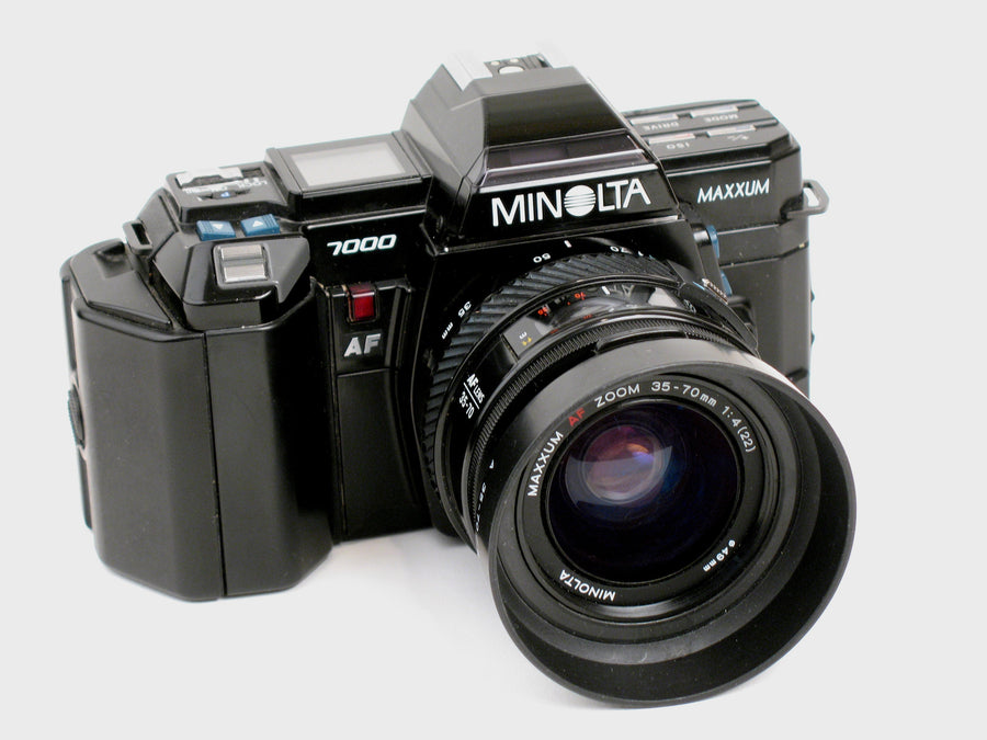 Black Minolta Maxxum 7000 SLR from 1985, seen from the front left, with the AF 35-70mm zoom lens and hood on a white background