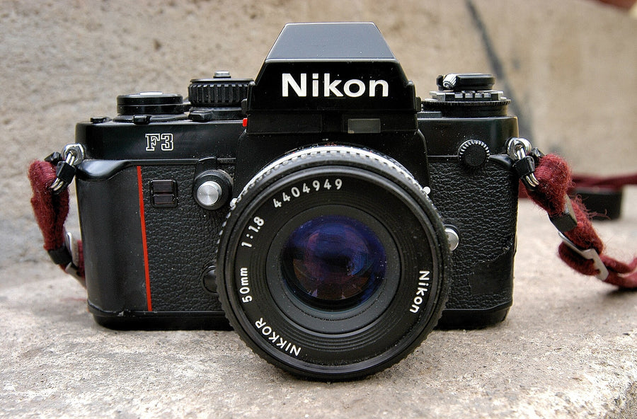 Black Nikon F3 seen from the front with a Nikkor 50mm f/1.8 lens and a burgundy strap, the thin red vertical line on the grip to the left of the lens