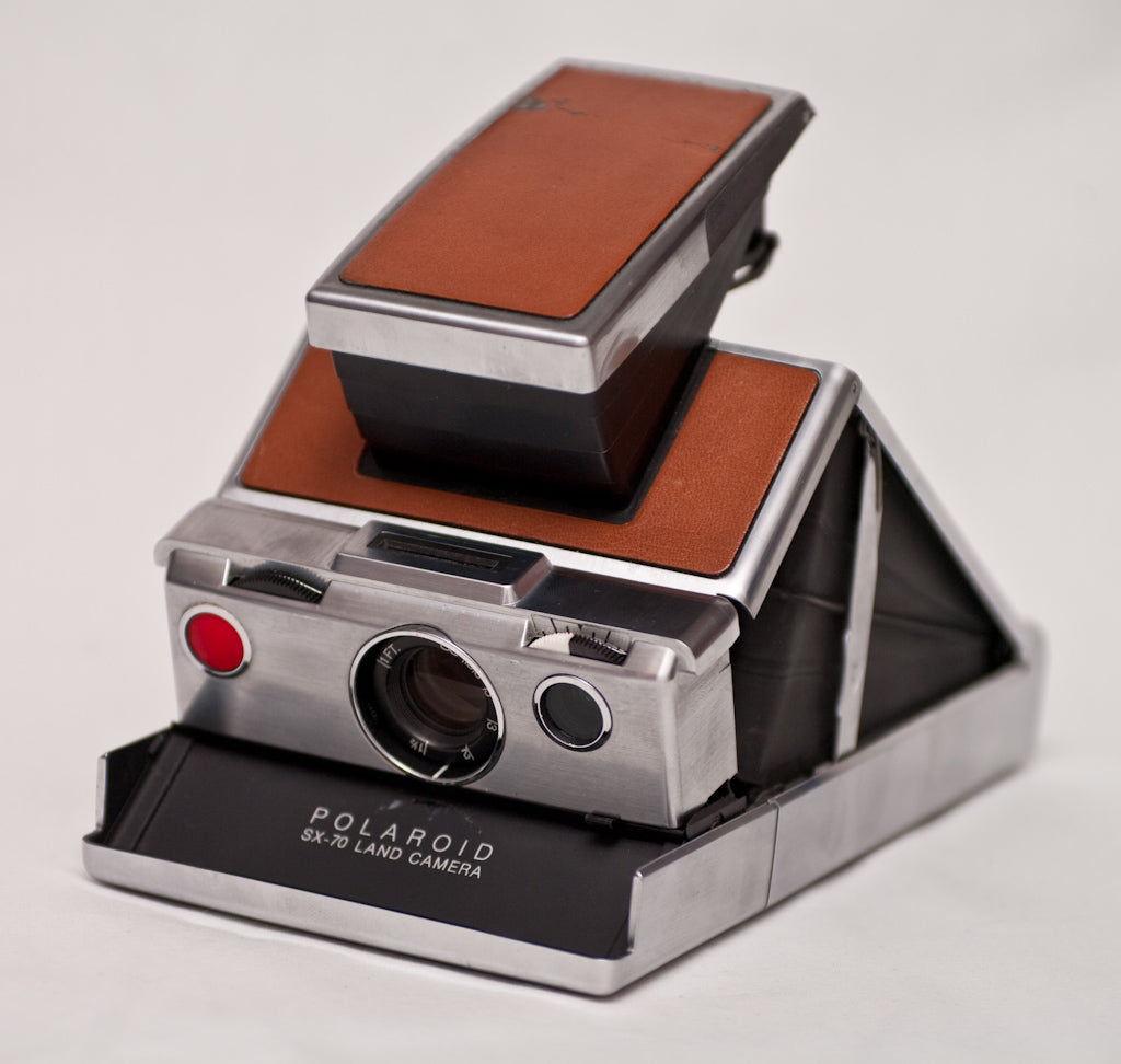 Polaroid SX-70 Land Camera opened, brushed chrome body with tan leather panels and red shutter button, seen from the front left on a light gray background